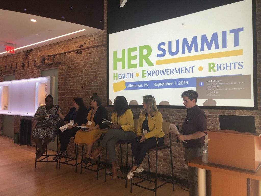 panel of speakers infront of ascreen saying "Her Summit"