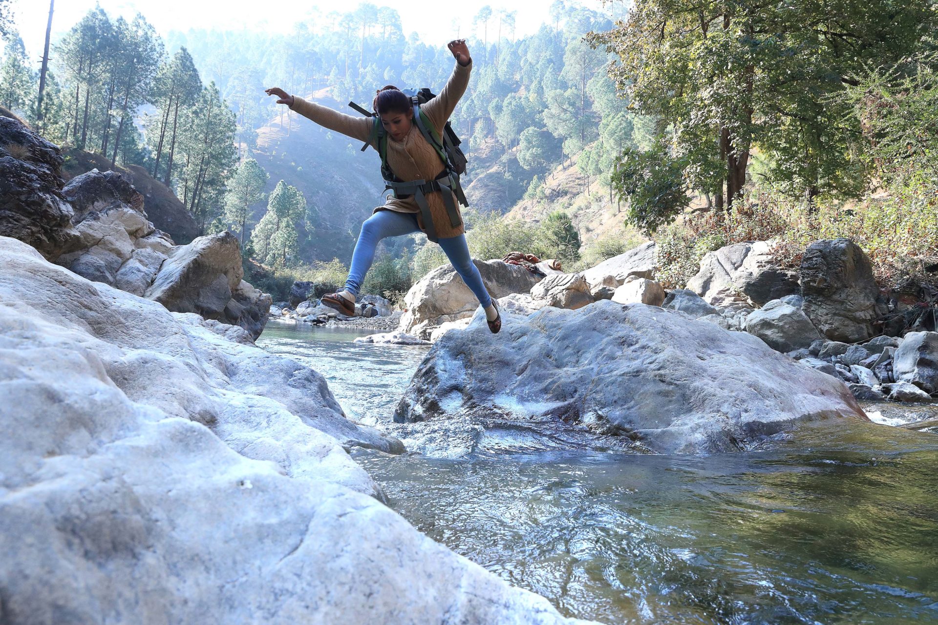 a Nepalese woman with her arms out in a U shape, one leg toward the rock she's aiming for and one leg in the direction from whence she jumped has a hiking backpack strapped to her back and is in mid-air of a jump across a fast moving river.