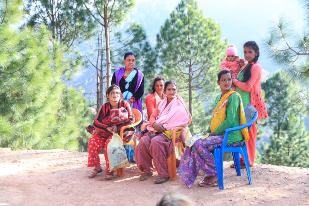 a group shot of at least 7 Nepalese woman in age range of newborn, toddler, and middle aged are all in colorful clothing like greens, purples, pinks and reds which are a stark contrast against rocky cliff edge they sit on and the woods that sit behind them