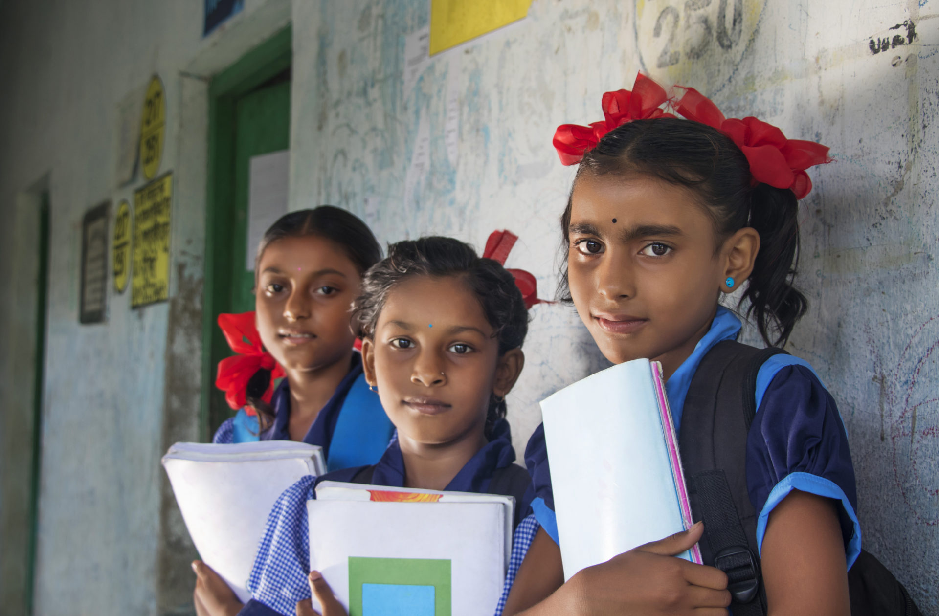 Three young female students stand outside of classroom holding books, rural India