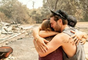 three people cry into a tight hug in front of the wildfire damage to their home.