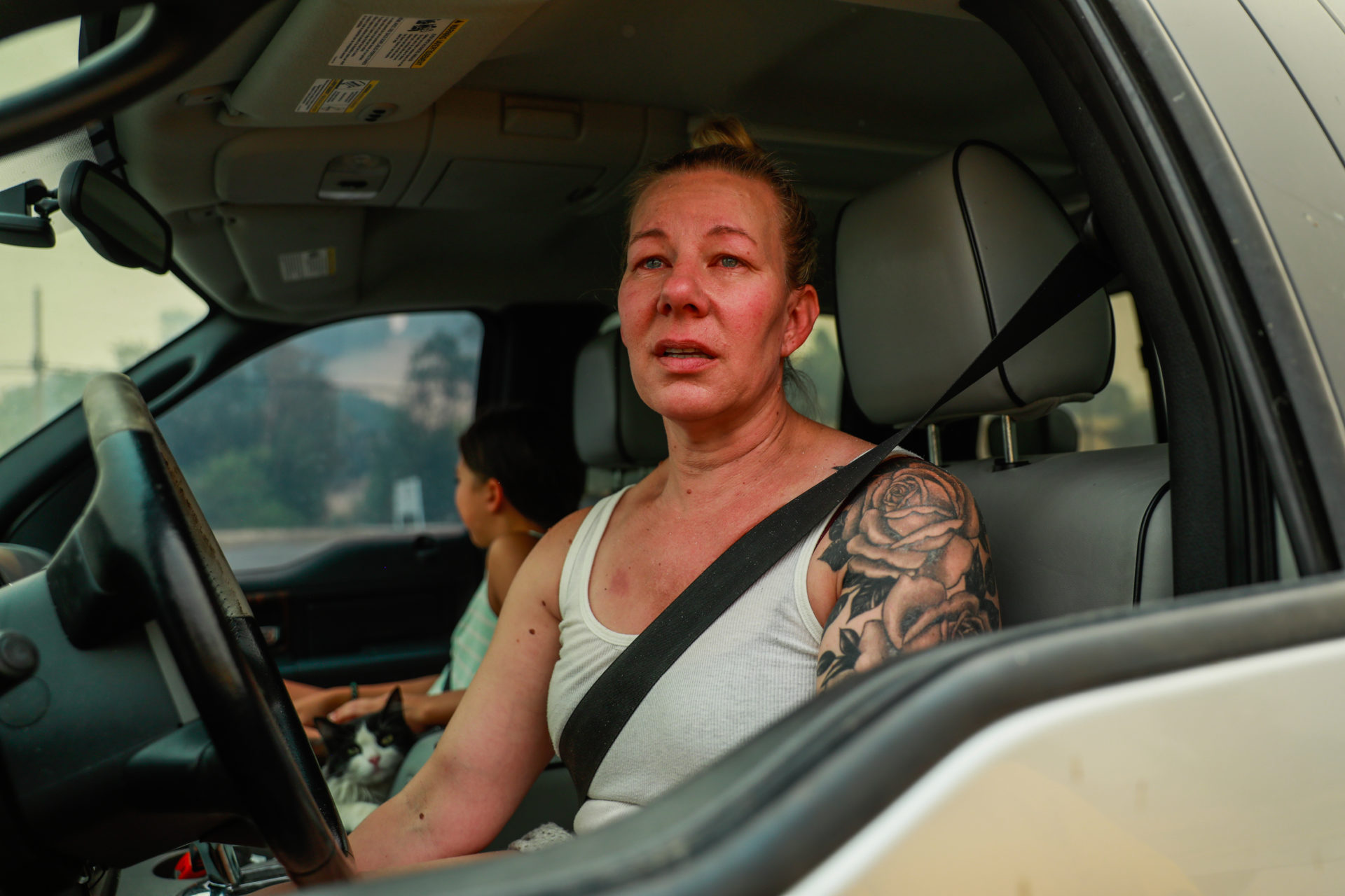 portrait of a white person in a white tank staring through the open car window onto the California wildfires, her face carrying the emotional weight of the sistuation.