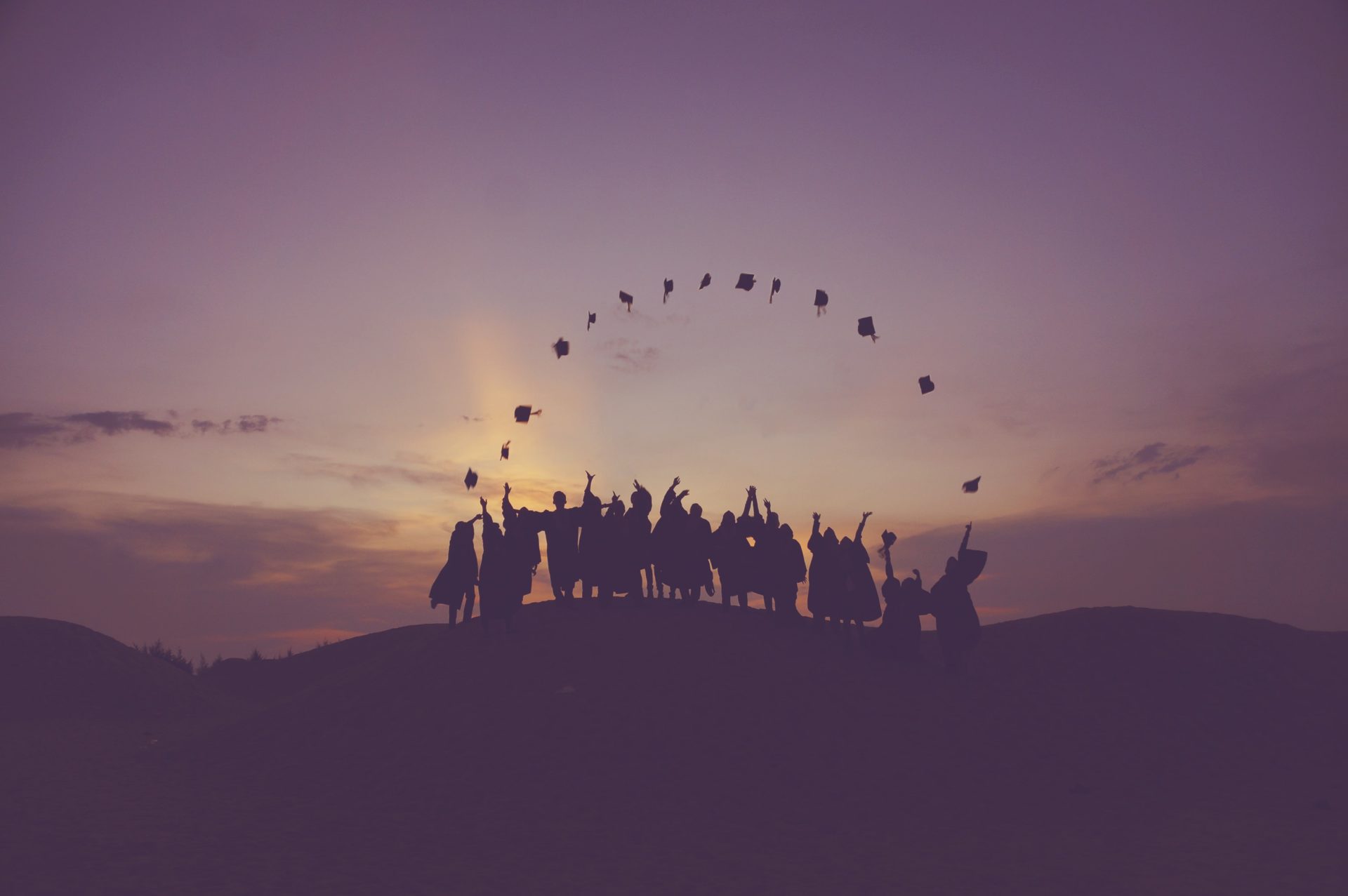 Group of recent graduates standing in a line together celebrating graduation by throwing their hats into the air. Hats make the shape of a half circle.