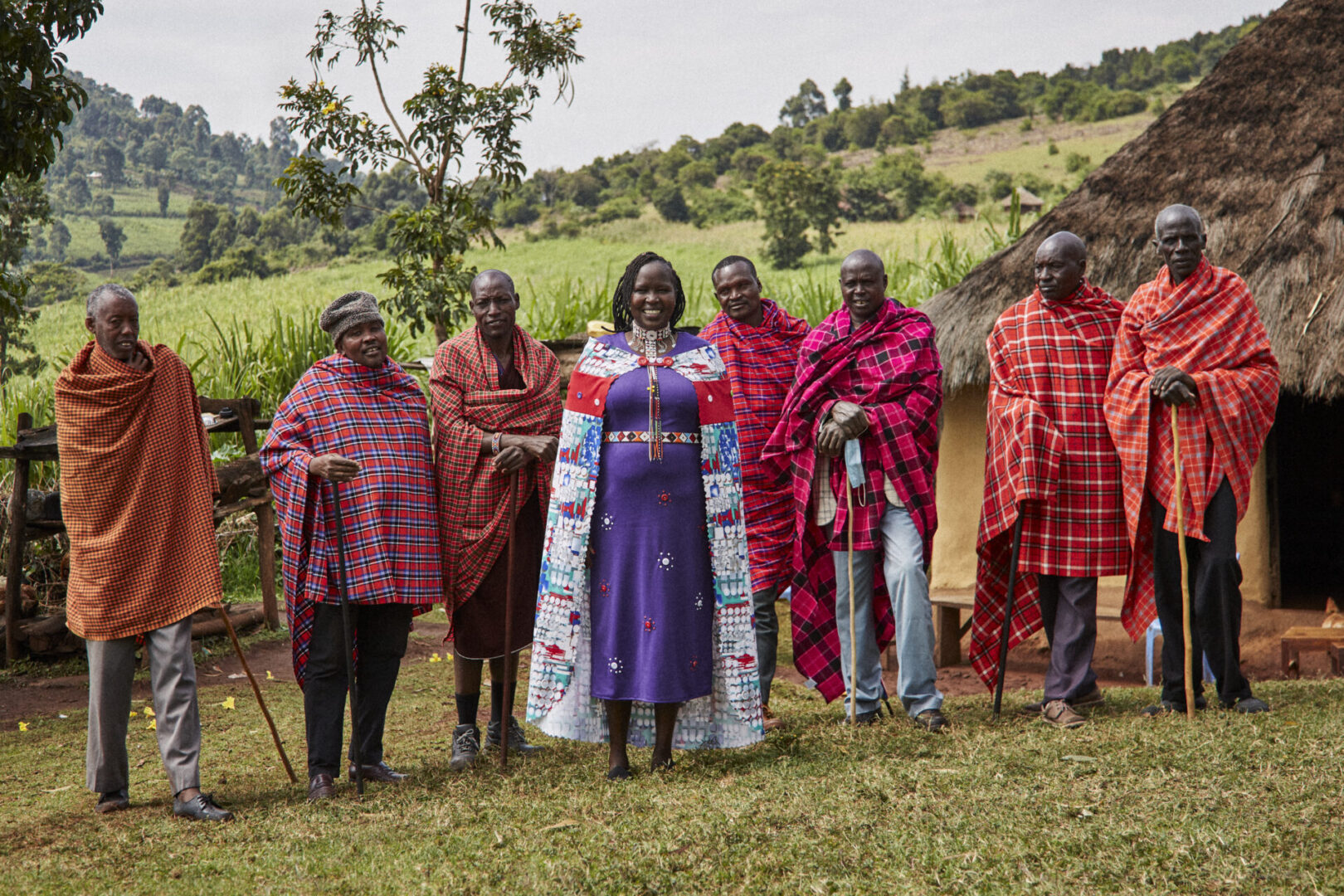 Dr. Kakenya Ntaiya stands in the middle of seven Maasai men, outdoors, in front of a Maasai home