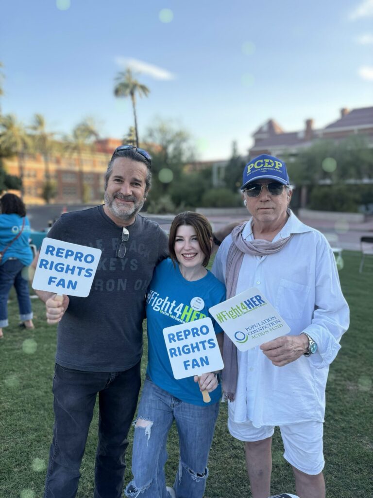 Kat Stratford stands in between two men; all three are holding paper fans that say "Repro Rights Fan"; Kat is wearing a blue #Fight4HER t-shirt