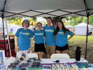 Four people (staff and volunteers) stand behind a table with branded giveaways, all wearing blue #Fight4HER t-shirts